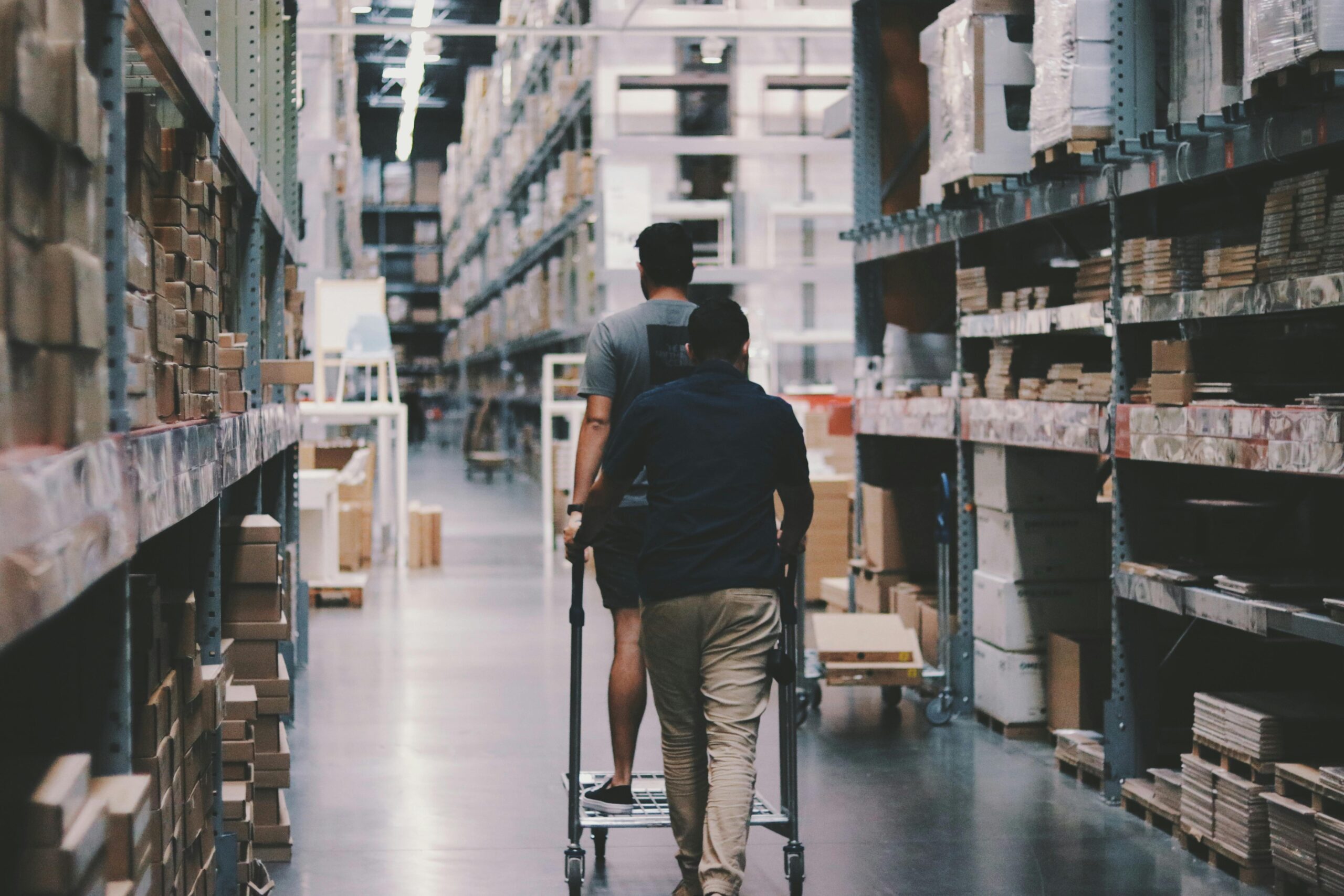 Two men maneuver a trolley in a large warehouse filled with boxes and shelves.